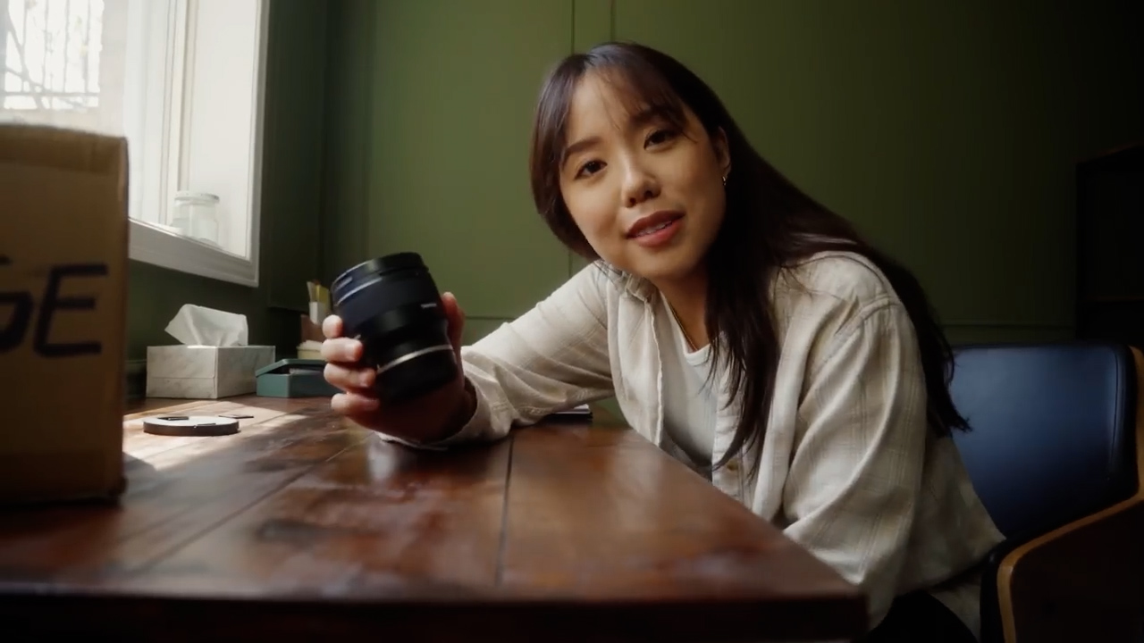 a young woman sits at a wooden desk, bathed in soft, natural light streaming in from a window.  she holds a camera lens, newly unboxed, with a gentle smile directed towards the viewer.  the scene exudes a cozy and inviting atmosphere, suggestive of a home studio or creative space.  behind her, a green wall adds a pop of color to the warm, earthy tones of the room.