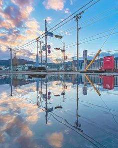 the image captures a serene and reflective street scene during sunset. the warm hues of the setting sun bathe the sky in a mix of orange, pink, and blue. power lines crisscross the top, leading the eye towards the vanishing point.  buildings and traffic signals line the street, their forms mirrored perfectly in a large puddle that dominates the foreground. the puddle creates a captivating symmetry, doubling the visual impact of the sky and urban elements. the overall feeling is peaceful and contemplative, showcasing the beauty found in everyday urban environments.