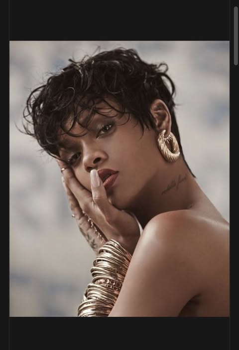the image is a portrait of rihanna. she has short, dark, wet hair and is wearing large gold hoop earrings and a stack of gold bracelets on her left arm. her left hand is resting on her face, showing off a manicure. she is wearing lipstick and has a tattoo on her neck. her shoulders are bare.