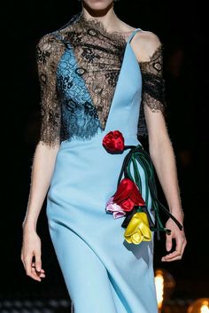 a woman is wearing a light blue dress with a black lace overlay. there are red, yellow, and green flowers on the front of the dress. she is posing on a runway with a dark background.