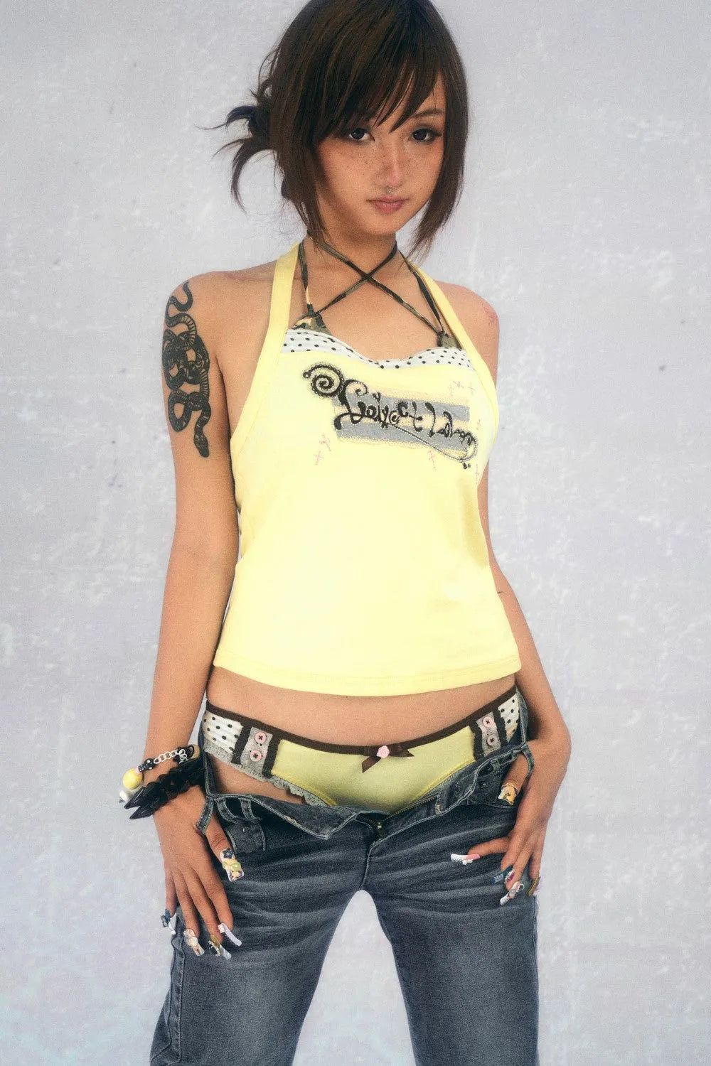 a young asian woman with short brown hair and bangs poses against a light grey background. she's wearing a pale yellow tank top with a graphic design, lowrise jeans, and brown and white patterned panties partially visible.  a snake tattoo is visible on her upper left arm. she also has long painted nails and several bracelets on her wrists. her expression is slightly serious, and she's looking directly at the camera. the overall style is casual and slightly edgy.