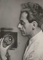 a black and white closeup portrait of a man, possibly a scientist or researcher, intently examining a camera lens or scientific instrument. he appears to be in a laboratory setting, with a partially visible piece of equipment in the background.  the image has a historical quality, suggesting it was taken sometime in the mid20th century. his expression is serious and concentrated, highlighting his focused attention on the object in his hands.  the overall mood is one of intellectual inquiry and scientific investigation.