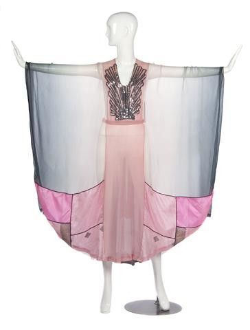 a mannequin stands displaying a vintage dress from the 1920s, a style often referred to as a flapper dress. the garment features art deco aesthetics with a sheer, sleeveless design. it is predominantly pink with black accents and is embellished with detailing around the vneck. the dress includes chiffon wings that extend from the shoulders and are black on the ends, and is a representation of the fashion of that era.