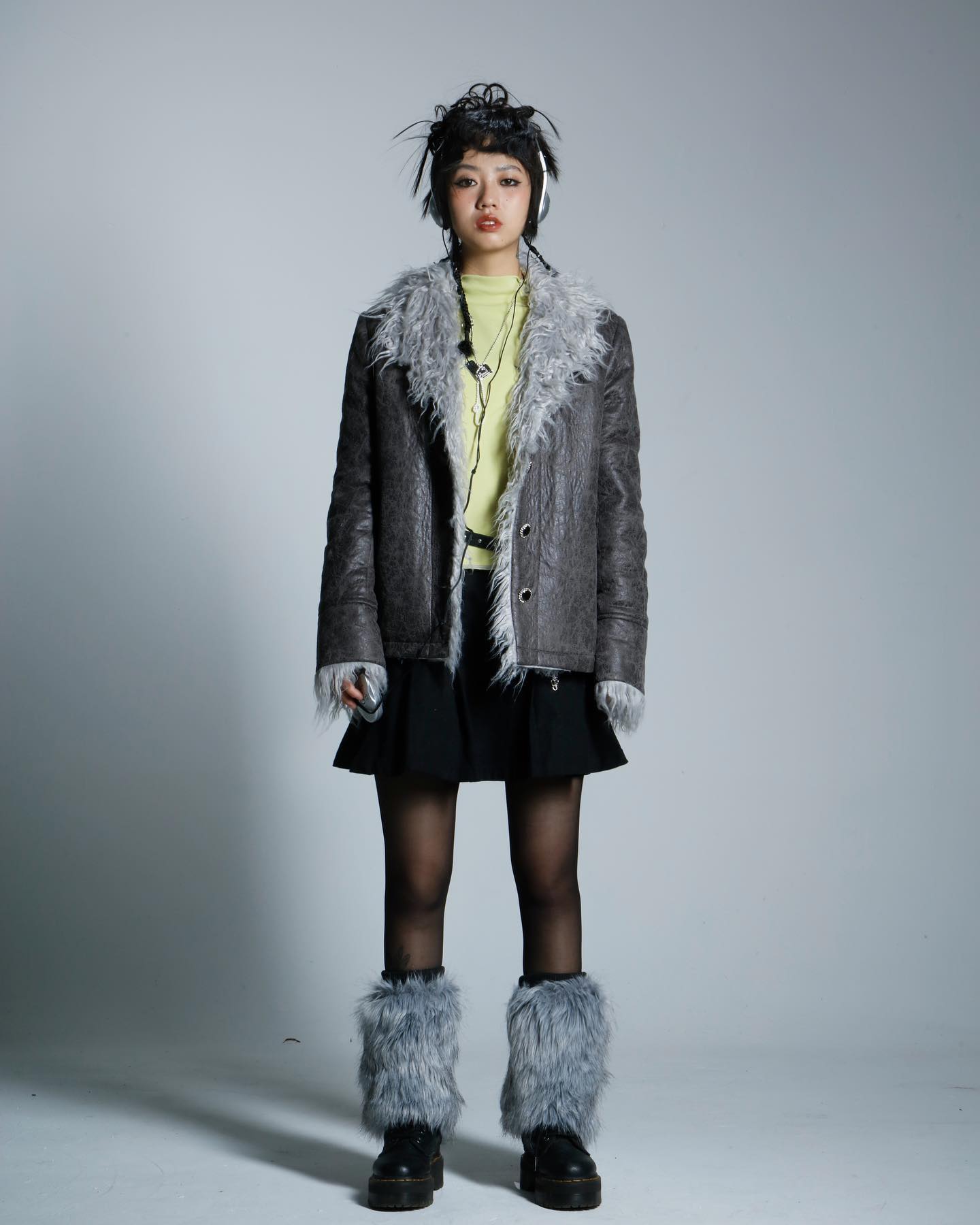 a fullbody shot of a young woman posing in a studio. she is wearing a grey faux furtrimmed jacket, a black mini skirt, black tights, grey fur leg warmers, and black boots. she also has headphones around her neck and her hair is styled in a slightly messy updo.  the background is plain grey. the overall style is edgy and fashionable.