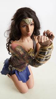 a woman in a wonder woman costume is kneeling on a white background. she is looking up at the camera with a determined expression. her hands are bound in front of her with what appears to be a ropelike material. she has detailed tattoos visible on her arm and shoulder.