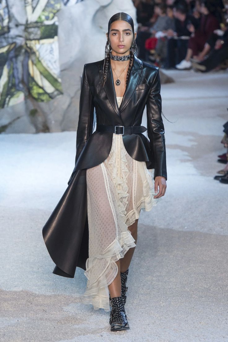 a female model walks the runway wearing a black leather jacket over a white ruffled dress. she also wears black studded boots. the model has her hair in braids and is wearing a silver necklace. the background is a grey and white runway.