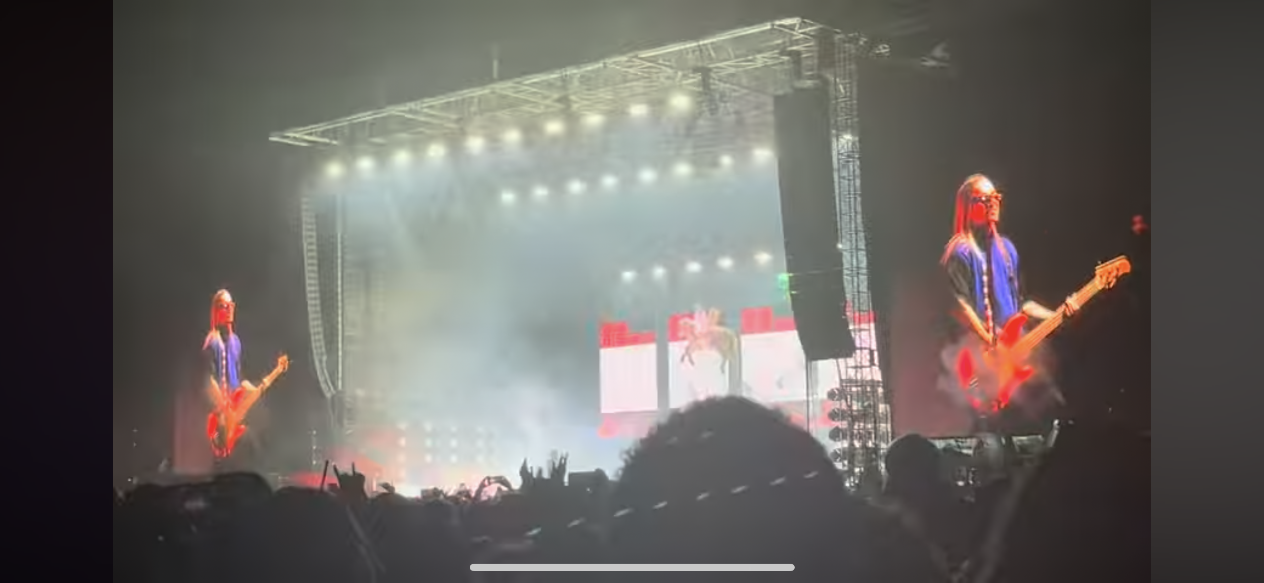 a view from the audience at a concert. the stage is lit up with bright lights and the band is playing in the background. the crowd is waving their hands in the air and enjoying the music. the guitarist is center stage and there is smoke and fog effects on stage.