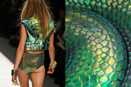 a fashion model walks the runway in a green, iridescent outfit with scales. the model's outfit is made of shiny, sequins and looks like snake scales.  the outfit is very glamorous and stylish.