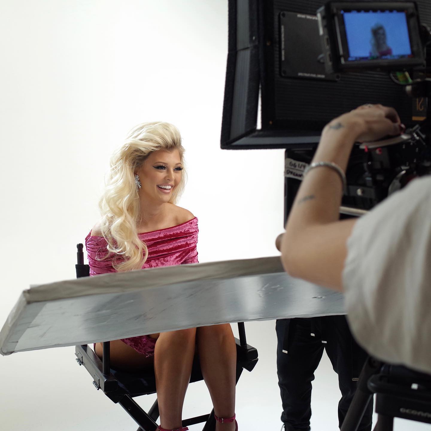 a woman with blond hair wearing a pink dress is sitting in a director's chair on a film set with a bright background. a camera and monitor are visible in the foreground, indicating the scene is being filmed behind the scenes.