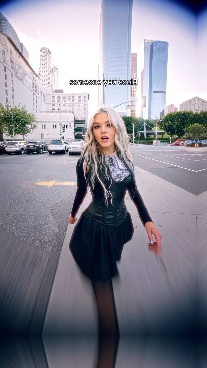 a woman with long white and blonde hair walks across a crosswalk in a city. she wears a black longsleeved top with a corset and a black skirt. several cars, buildings and skyscrapers can be seen around the image. there is text on the image that says "someone you could".