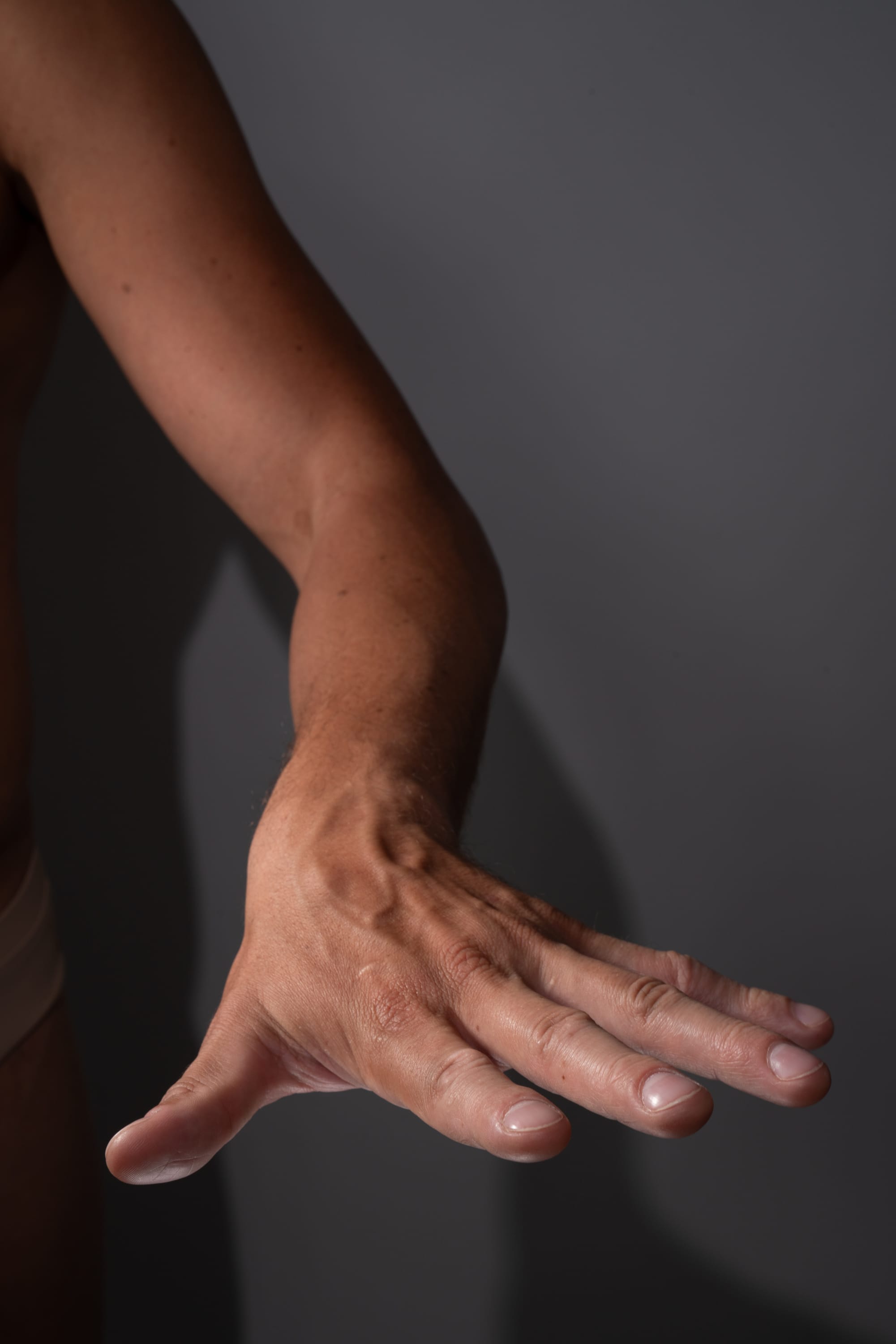 a closeup view of a man's outstretched arm and hand, showcasing the anatomy and details of the skin and veins under studio lighting. the arm extends towards the viewer, fingers slightly splayed, against a neutral gray background.