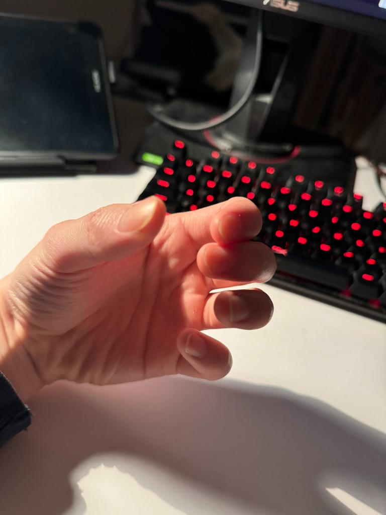 a left hand is shown with fingers slightly curled. in the background is a black keyboard with red glowing keys. a black computer monitor and black tablet are out of focus in the background.