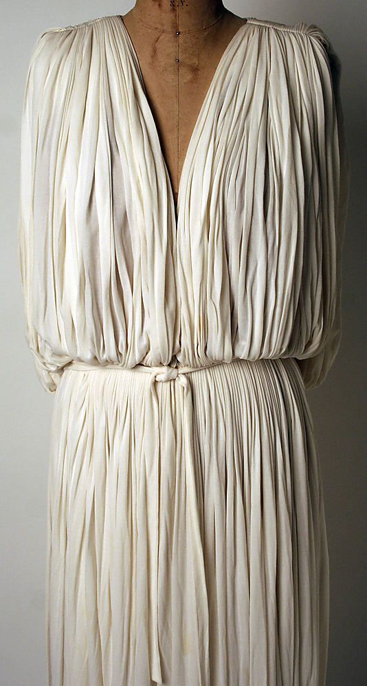 loseup view of a creamywhite pleated dress, possibly inspired by ancient greek or roman styles. the dress is displayed on a dress form, showcasing its fabric texture and construction details. the pleats are meticulously formed, and a simple tied belt cinches the waist. the fabric drapes softly, highlighting the elegance and simplicity of the design. the background is a muted, neutral color, ensuring the dress is the main focus of the image. the image is sharp, allowing for a clear view of the fine details of the dress's fabric and construction.