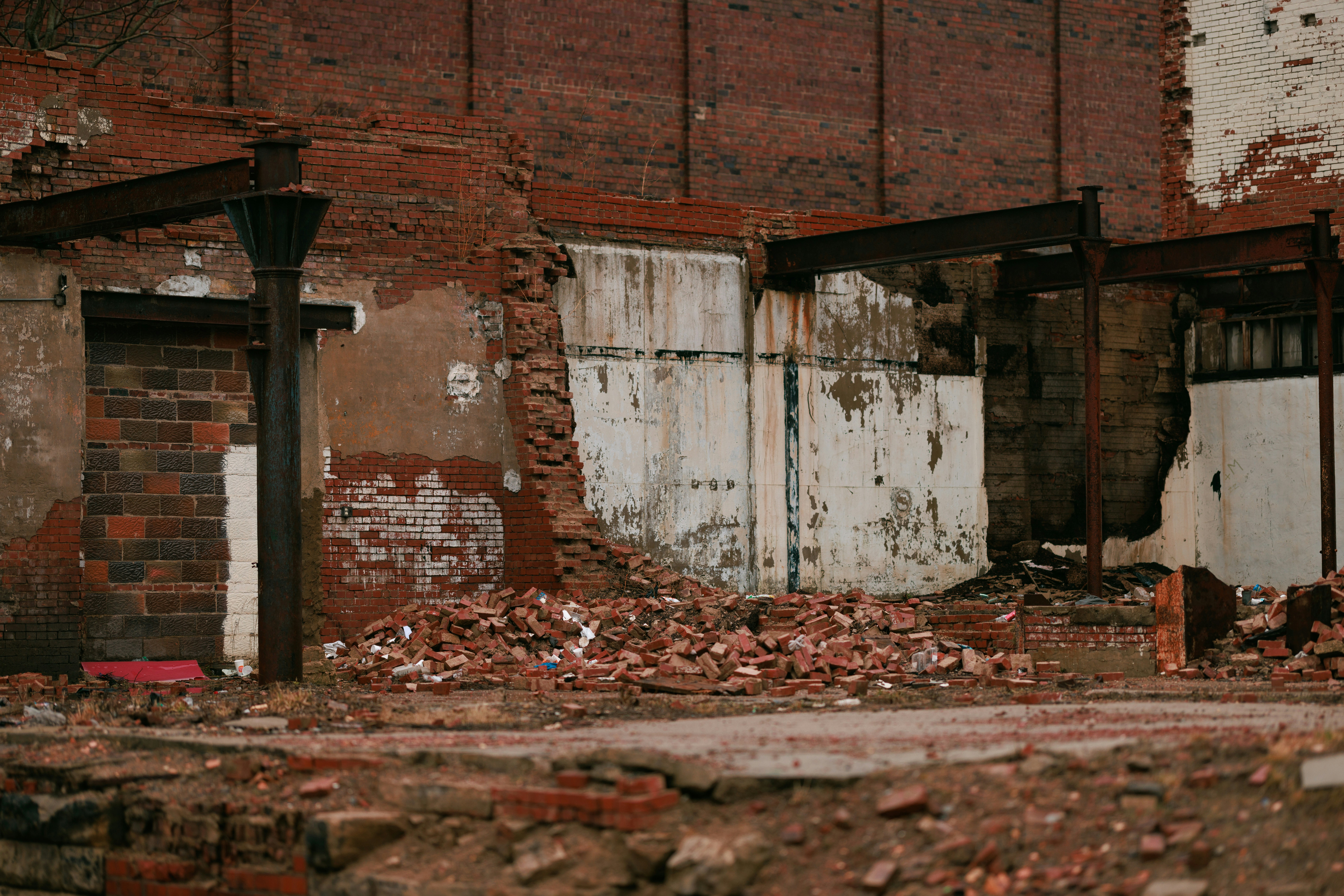 the image shows the remnants of a demolished brick building. the walls are partially standing, revealing a pile of rubble inside.  rusty metal beams protrude from the walls, indicating a former structure. the ground is covered with bricks, dirt, and debris.  the overall color palette is muted and somber.