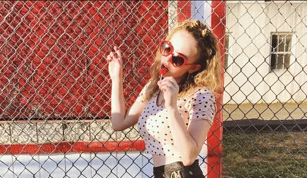 a young blonde woman with curly hair is posing playfully in front of a red chainlink fence. she's wearing red round sunglasses, a pink polka dot crop top, and dark shorts. she's holding a red lollipop and seems cheerful and happy. the setting appears to be outdoors on a sunny day.