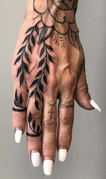 a hand with multiple tattoos is shown in a closeup shot. the tattoos include a flower on the back of the hand, vines with leaves extending down the hand and onto the fingers, a small cross on one finger, and an infinity symbol on another. the person is wearing white nail polish and what appears to be a ring tattoo around one finger. the background is a plain gray.
