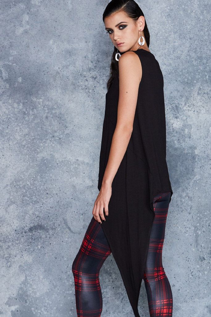 a woman is posing in a studio. she has dark hair and is wearing a black sleeveless dress and red plaid leggings. she is wearing large earrings and looking at the camera. the background is gray.