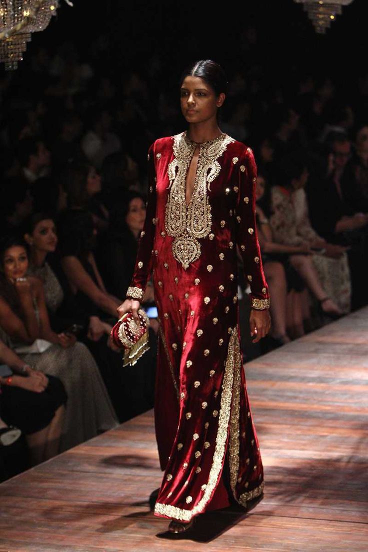 the image shows a runway model gracefully walking down a wooden catwalk. she is wearing a stunning floorlength anarkalistyle gown in rich red velvet, lavishly embellished with intricate gold embroidery.  the dress features a high neck, a deep vneckline, and side slits. the model carries a small, ornate clutch. the background is blurred, showcasing a seated audience watching the fashion show.  the overall ambiance suggests a highfashion indian clothing event.