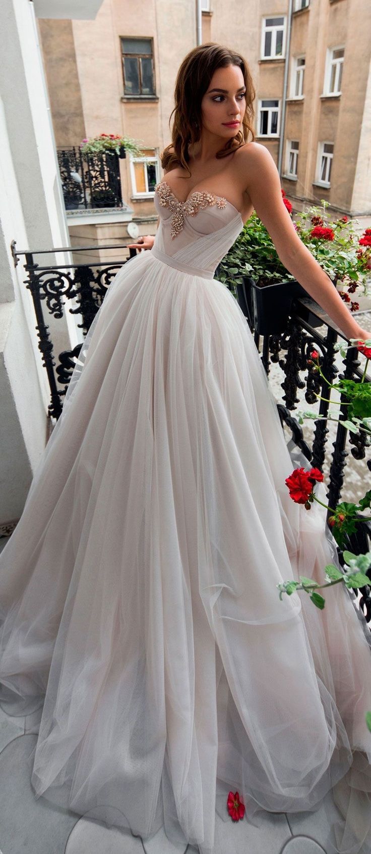 a woman in a white wedding dress is standing on a balcony. the dress is aline with a sweetheart neckline and is made of tulle. the balcony is decorated with flowers. the woman is looking to the right of the frame. the lighting is natural daylight. the image is taken from eye level.