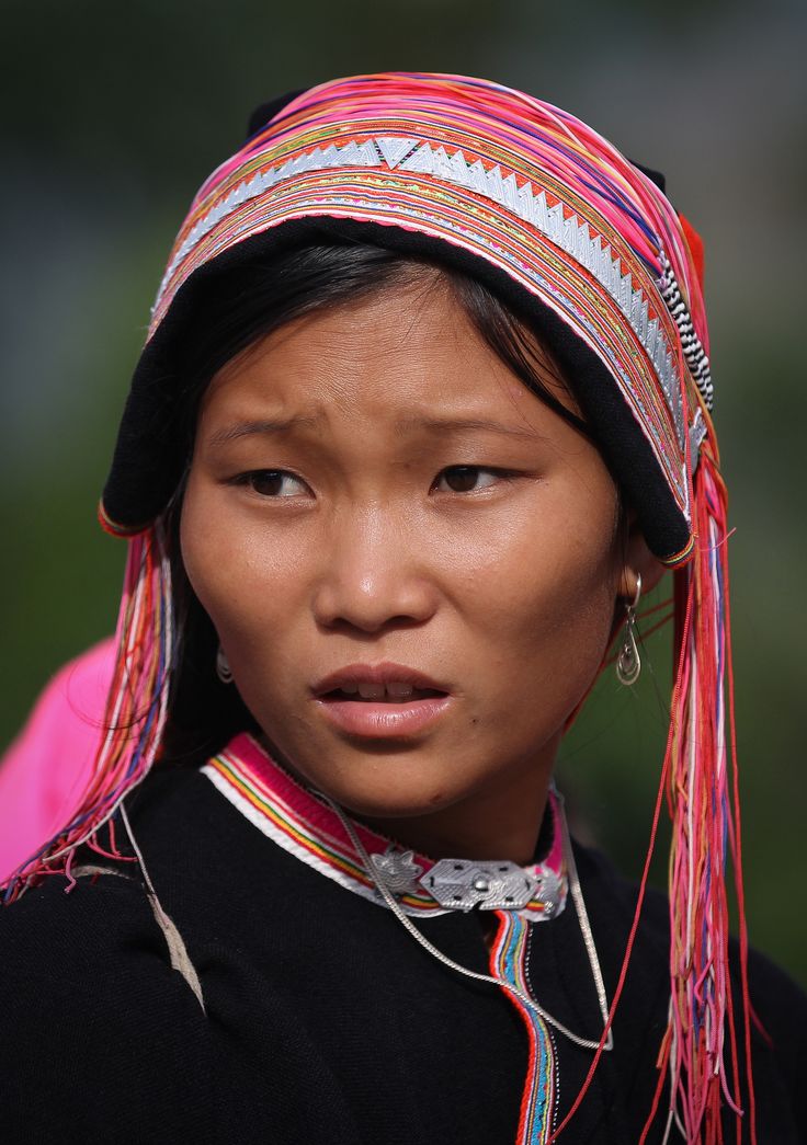 a young asian woman in traditional clothing looking at the camera. she is wearing a black outfit with a colorful tribal hat with fringe and earrings.