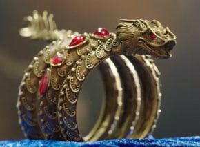 a closeup shot showcases a golden dragon bracelet adorned with red gemstones, resting on a blue cloth. the bracelet features intricate scales and a detailed dragon head.
