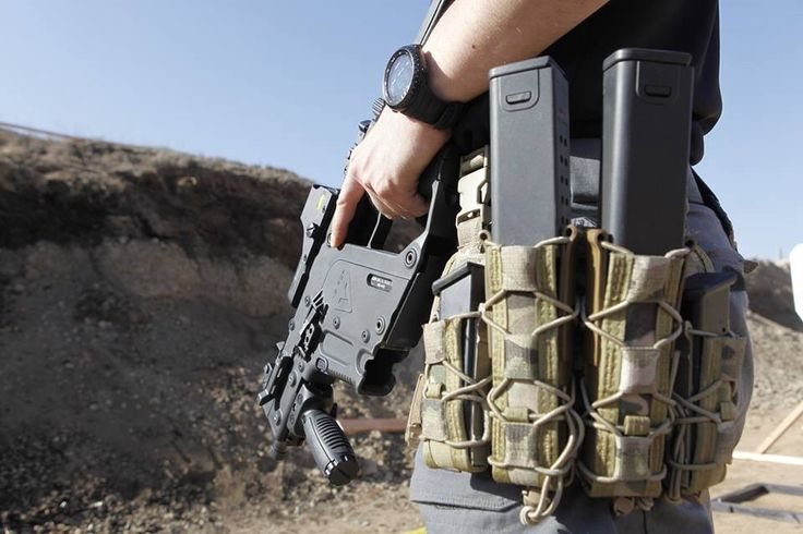 a person is holding a handgun with a tactical belt and military camouflage magazine pouches. they are wearing a black watch and the background shows a dirt hill.