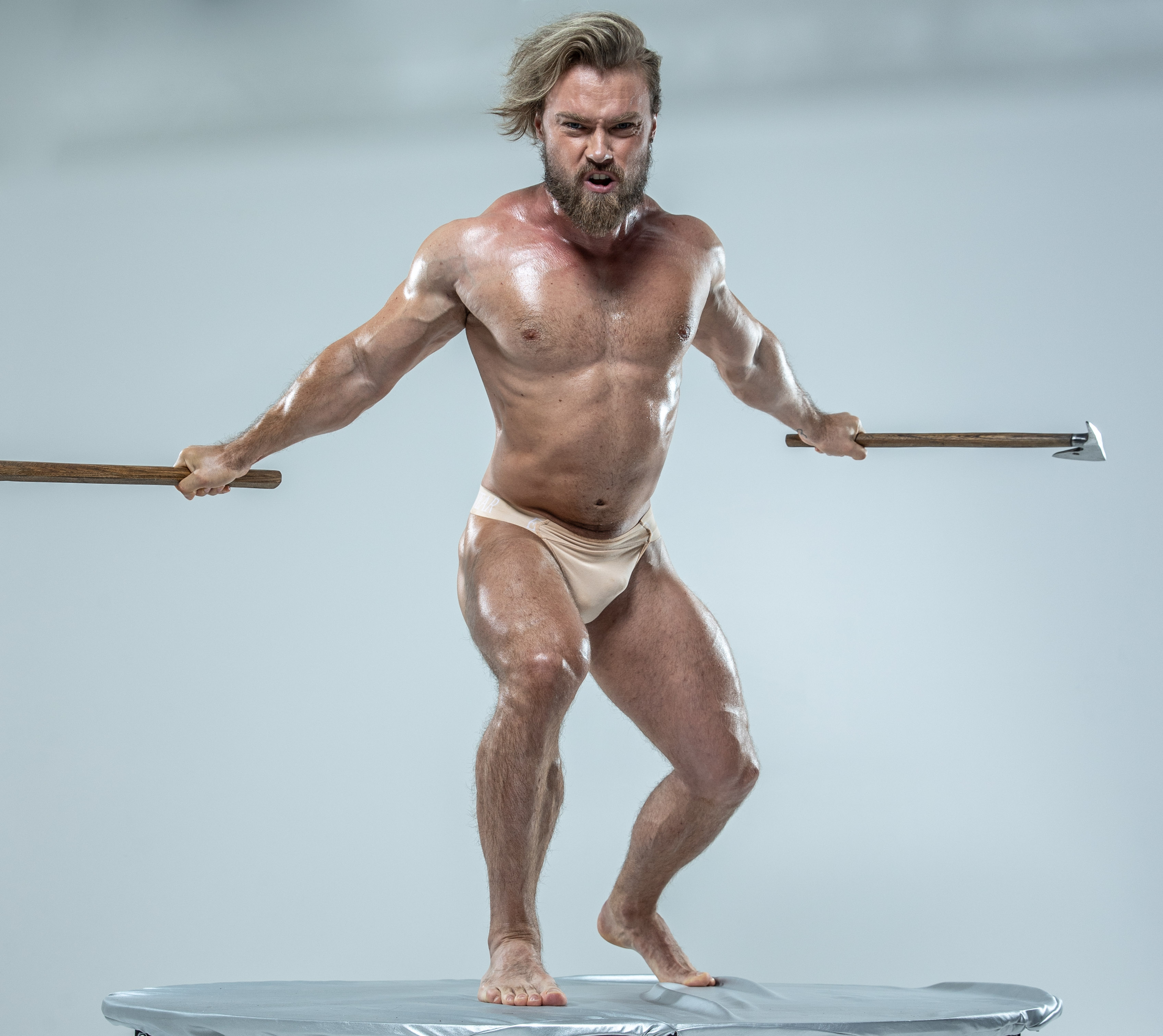 a muscular man with long blond hair and a beard stands barefoot on a round, metallic platform against a light gray background. he is shirtless, wearing only a pair of white underwear, and his body is glistening with sweat.  the man is posed dynamically, legs apart and slightly bent, as if in midbattle. he holds a wooden axe in each hand, raised aggressively, with a determined and fierce expression on his face. the overall image evokes themes of strength, aggression, and possibly norse or barbarian mythology. the studio lighting is bright and highlights the man's muscular physique.