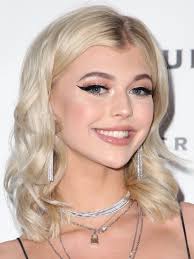 a portrait of loren gray smiling with blonde, shoulderlength wavy hair, wearing eyeliner and multiple layered necklaces.