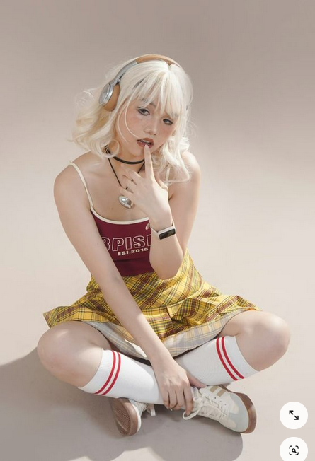a young woman with blonde hair is sitting on a floor wearing headphones. she is wearing a red tank top with a yellow plaid skirt and white socks with red stripes. she
