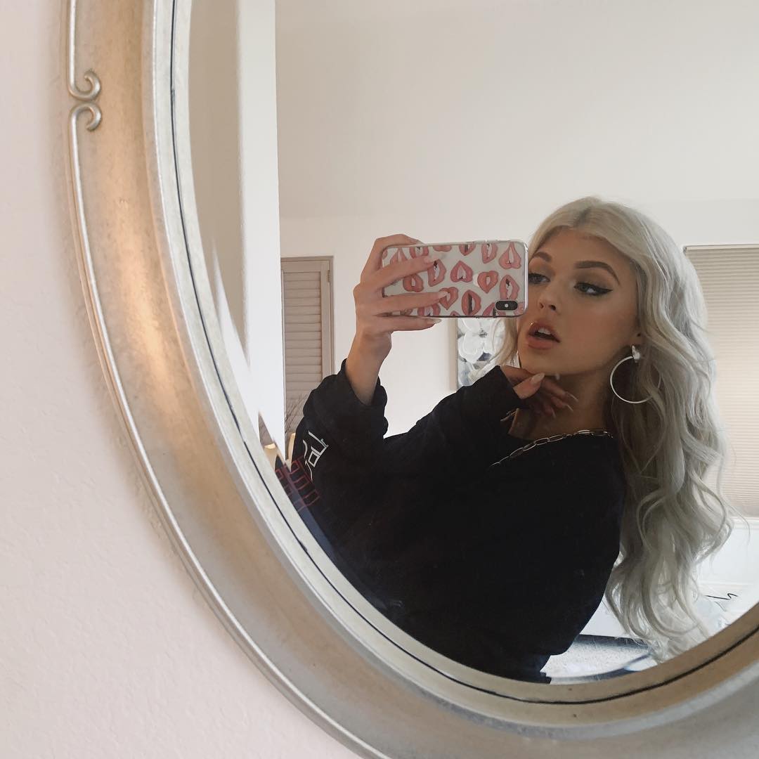 a woman with long, silver hair and wearing a black sweater is taking a selfie in front of a round, silverframed mirror. she is holding a phone with a white case covered in red lip prints. she has on makeup, including lipstick and eyeliner, and is wearing silver hoop earrings and a silver chain. the background shows a bedroom setting with blinds and a door.
