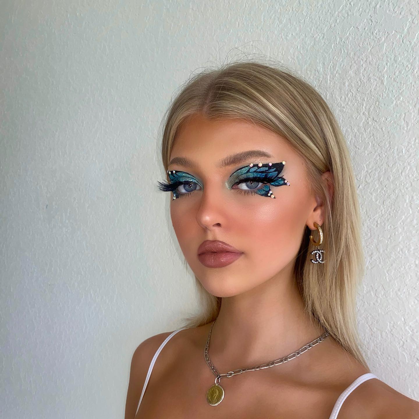 a portrait of a woman with blonde hair and blue eyes, wearing dramatic butterflyinspired eye makeup. she is also wearing jewelry, including chanel earrings and a coin necklace, and a simple white tank top.