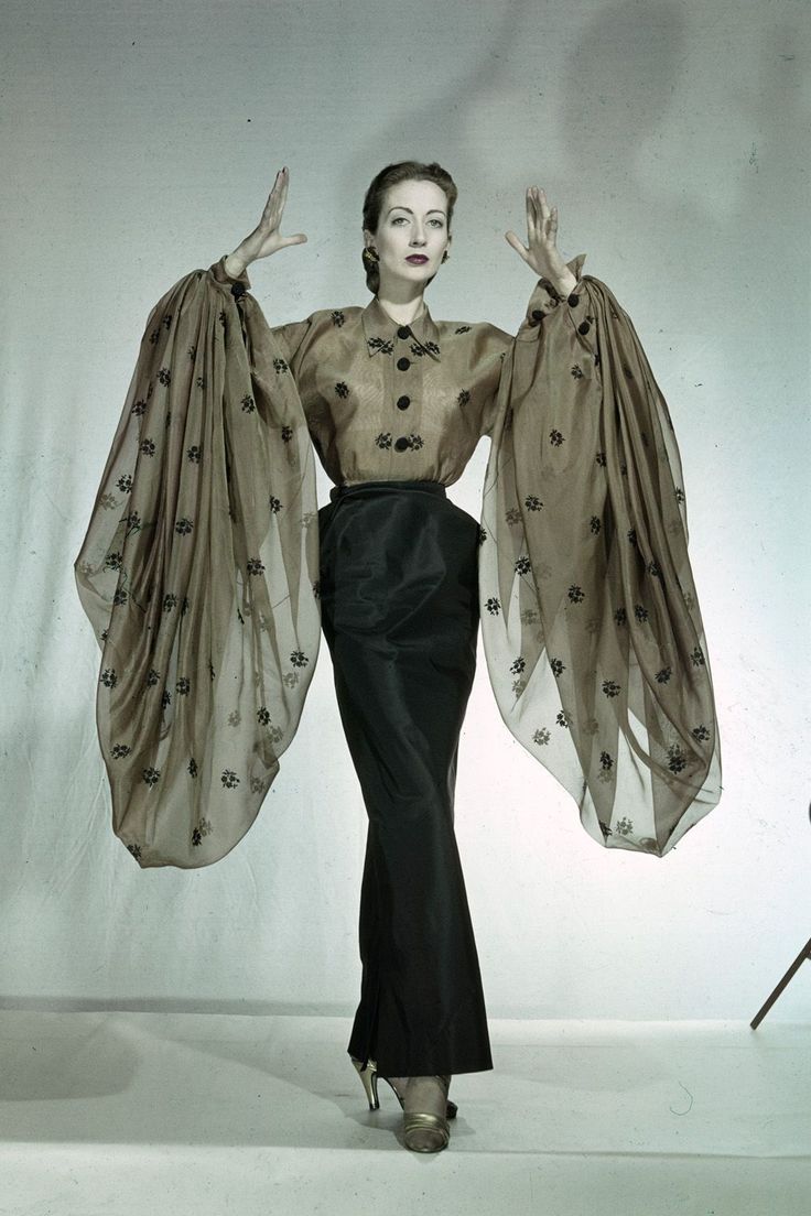 a vintage color photograph depicts a woman in a striking fashion ensemble, standing with her arms raised and palms facing forward against a neutral background. she wears a sheer, taupecolored blouse adorned with small, dark floral or abstract motifs. the blouse features a row of dark buttons down the front and dramatically oversized, billowing sleeves that extend far beyond her hands. the blouse is tucked into a highwaisted, black skirt that is fitted through the hips and thighs, tapering slightly towards the bottom. she is wearing high heels that match the blouse in color, and her hair is styled in an elegant updo. her makeup includes dark lipstick and defined eyebrows. the overall impression is one of sophisticated elegance and dramatic flair, typical of mid20th century high fashion. a partial leg of a photography tripod is visible in the background on the right.