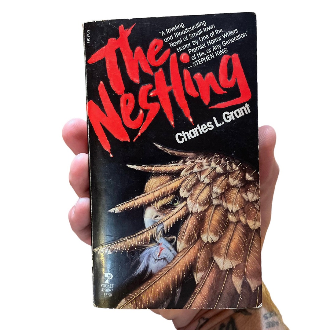 a closeup shot of a person's hand holding a vintage paperback copy of "the nestling" by charles l. grant. the book cover features an illustration of a large bird of prey, possibly an eagle, with its feathers prominently displayed.  a quote from stephen king praising the book is visible on the cover. the book's spine shows it is a pocket books publication, with the price of $3.50. the overall condition of the book suggests it's a used copy.