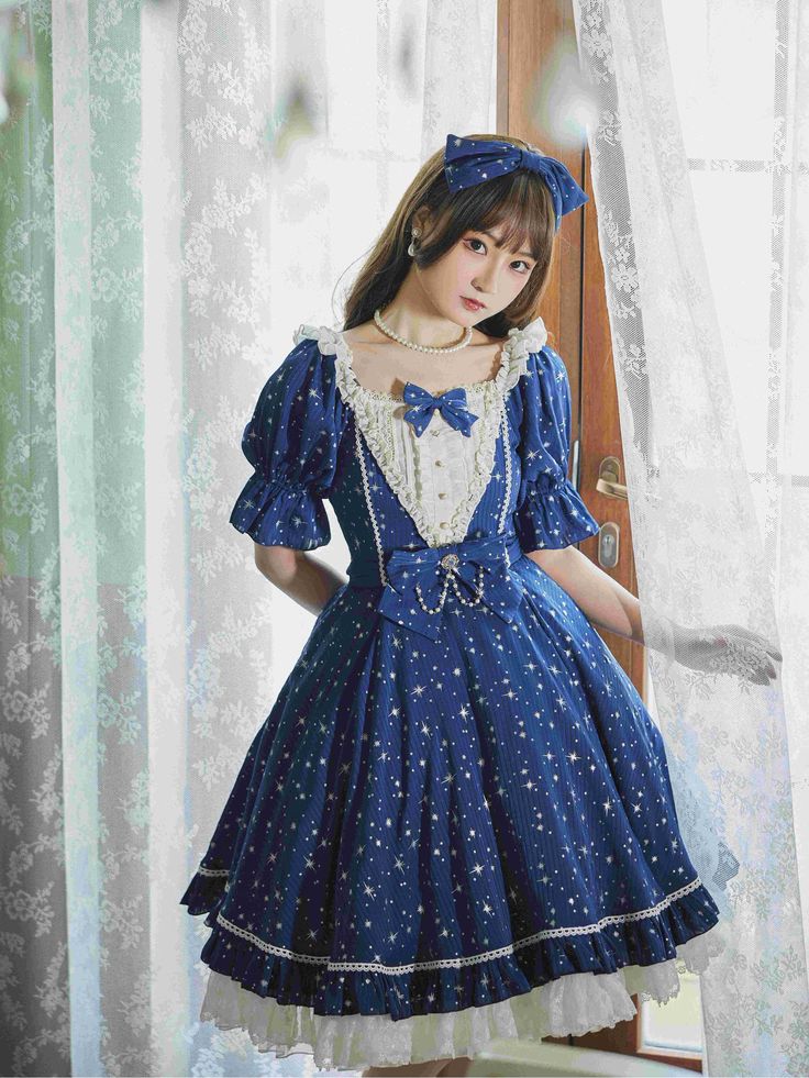 a young woman poses in a blue and white lolitastyle dress. the dress features a starry pattern, puff sleeves, and lace detailing. she is standing in front of a window, which provides soft, natural lighting.