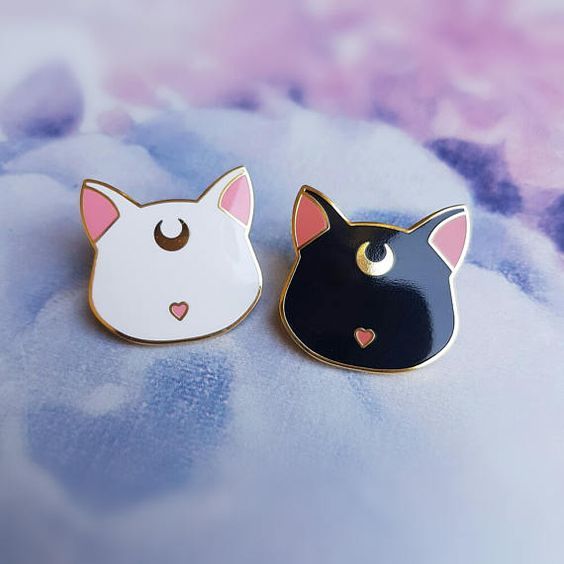 the image shows two enamel pins of cat faces, one white and one black, with gold outlines, pink inner ears, crescent moon symbols on their foreheads, and heartshaped noses. they are placed on a surface with a soft, watercolorlike floral pattern.