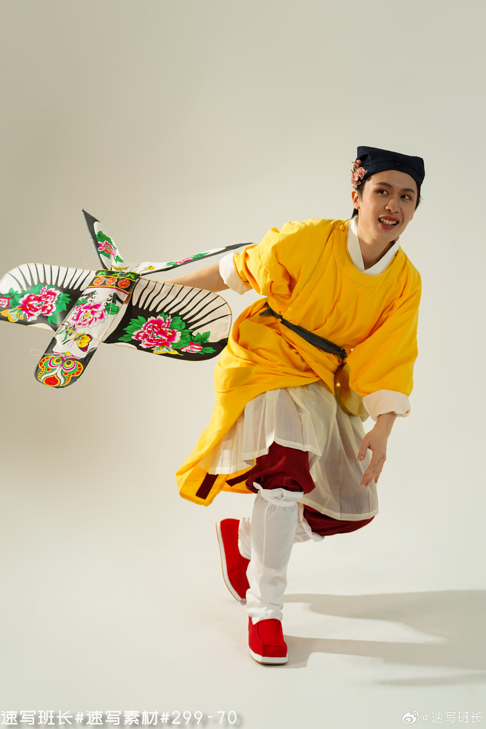 a young man wearing a vibrant yellow traditional chinese robe and red shoes is captured midmotion, appearing to run and joyfully fly a colorful kite decorated with floral and bird motifs. the background is a clean, bright white, suggesting a studio setting with controlled lighting. the image emphasizes the dynamic movement and vibrant colors of the scene.