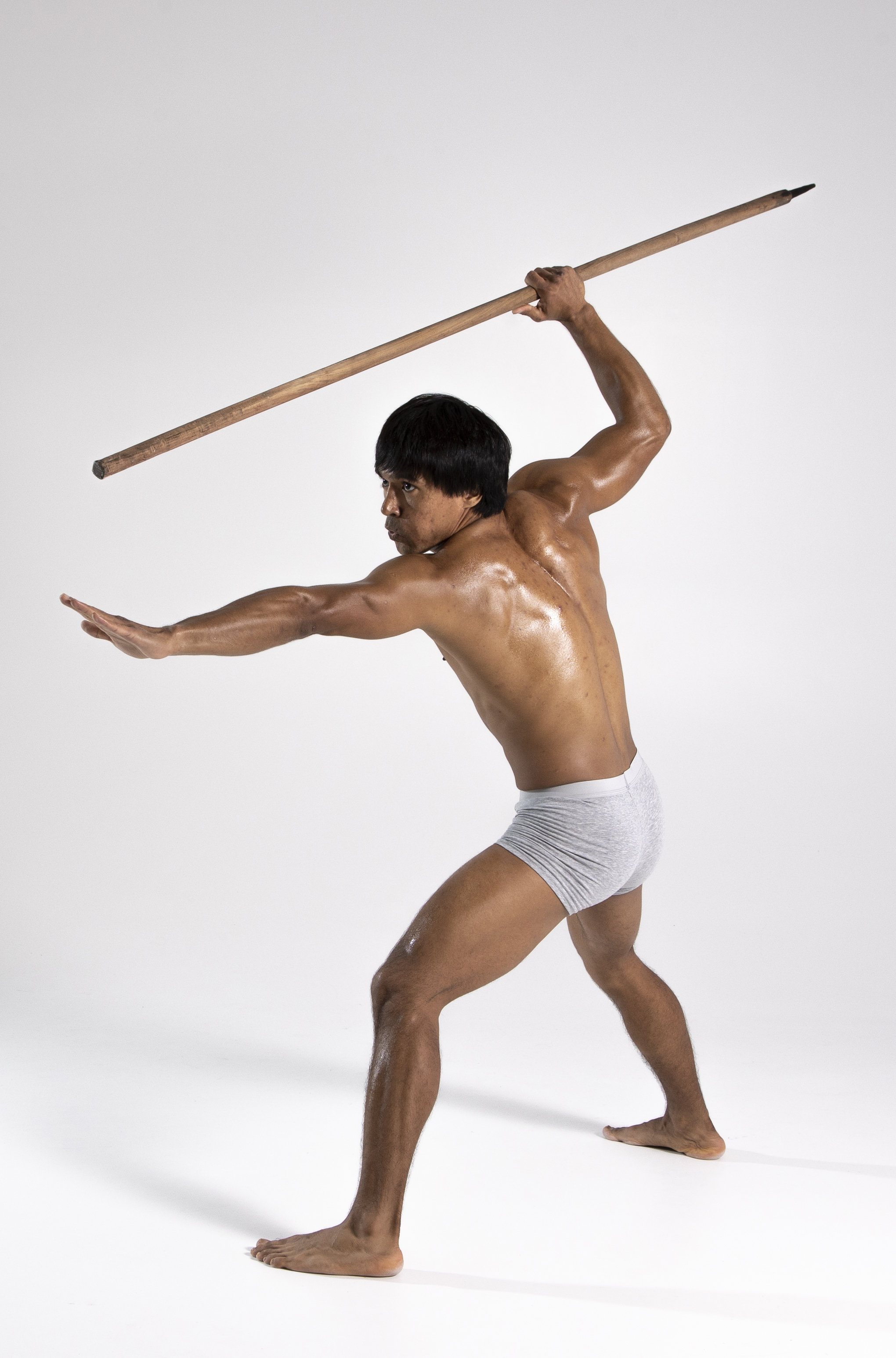 a shirtless, muscular man wearing a loincloth poses in a warrior stance, wielding a spear in a studio against a white background. he is looking to the side, with one arm extended and the spear held above his head, ready to attack.