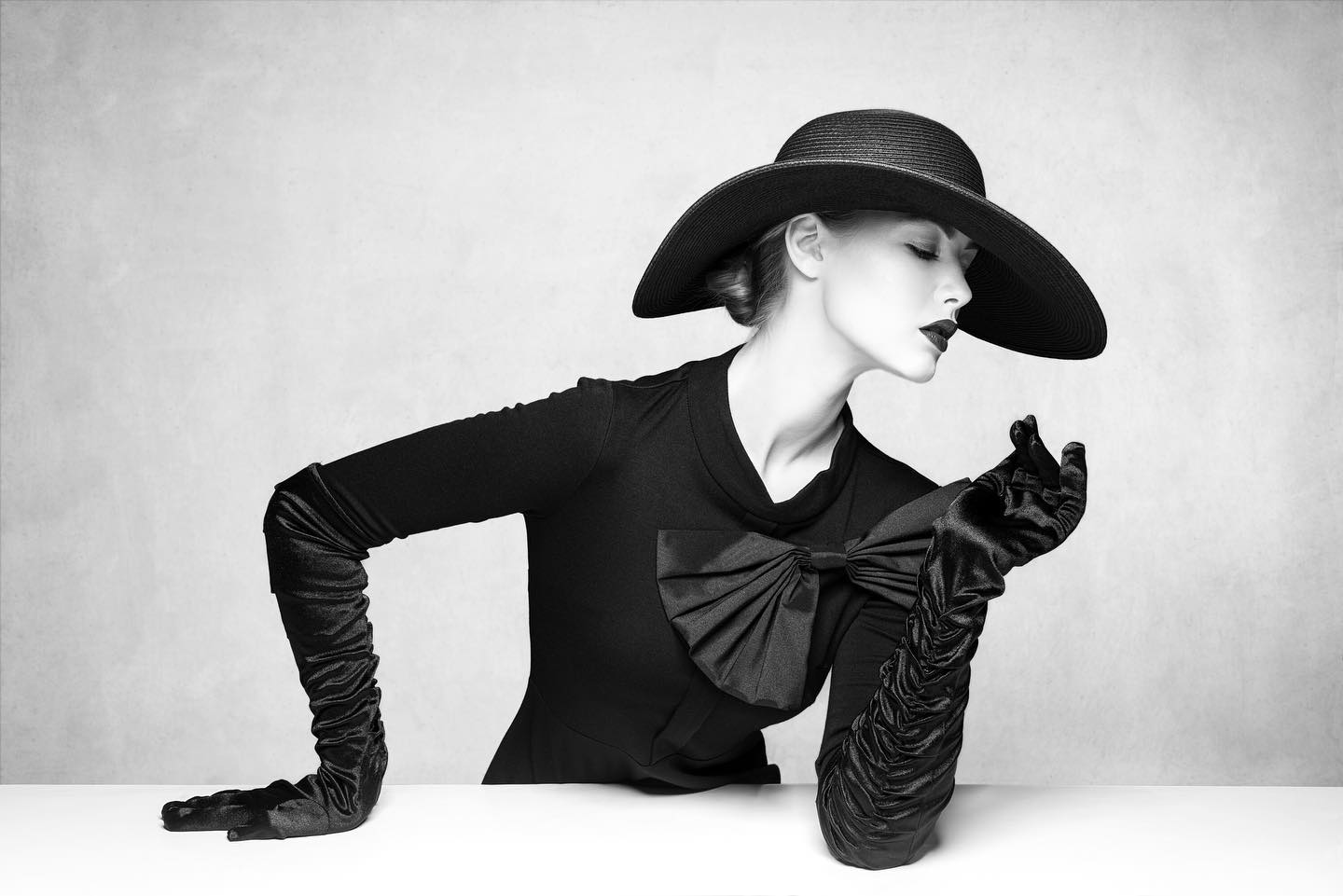 a black and white portrait of a woman in a black hat and gloves. she is wearing a black dress and has a serious expression on her face. she is looking down and her pose is dramatic and mysterious. she is standing in front of a plain white background and her black outfit stands out against the white. she has a large bow on her dress. the image has a vintage feel.