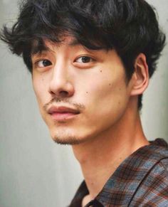 a closeup portrait of a young asian man with dark hair and a light brown plaid shirt. he has a mustache and goatee, and his dark eyes are looking directly at the camera. the background is a light gray wall, and the image is lit with soft lighting.