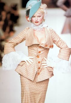 a blonde woman in a retro plaid dress and hat walks down a runway. she is wearing white lace gloves and has a chic, elegant style.