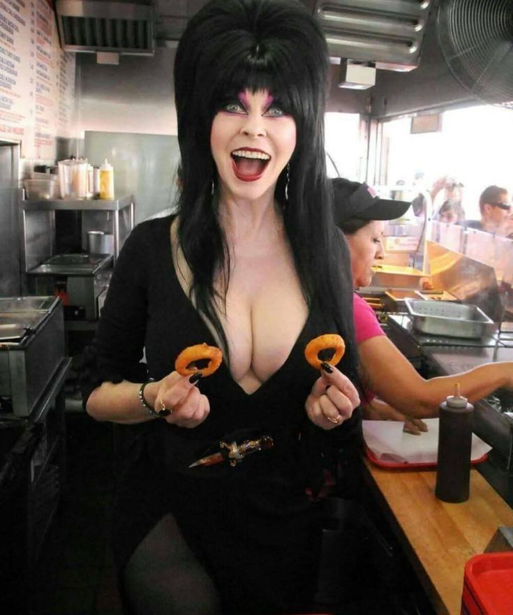 a woman with long black hair and a black dress is holding two onion rings and smiling. she is standing at a counter in a restaurant.