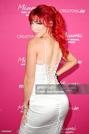 a woman with vibrant red hair is captured in a side and back profile shot. she's elegantly dressed in a formfitting white dress that features a unique lacedup back detail. the setting appears to be an event, suggested by the pink background adorned with "creators miami" branding. the overall mood is glamorous and stylish.