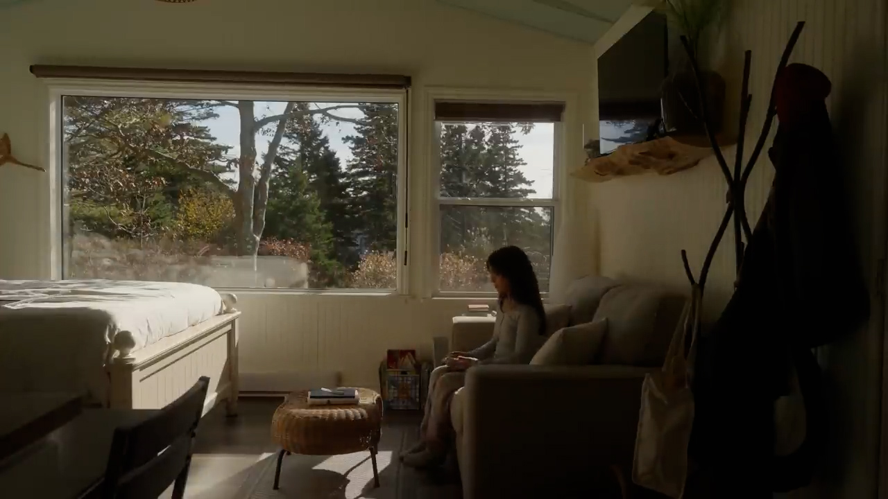 a woman is sitting on a couch in a cabin, reading a magazine. the cabin has large windows with a view of trees and the ocean. the room is bright and airy, with natural light streaming in through the windows. the overall atmosphere is one of peace and relaxation.