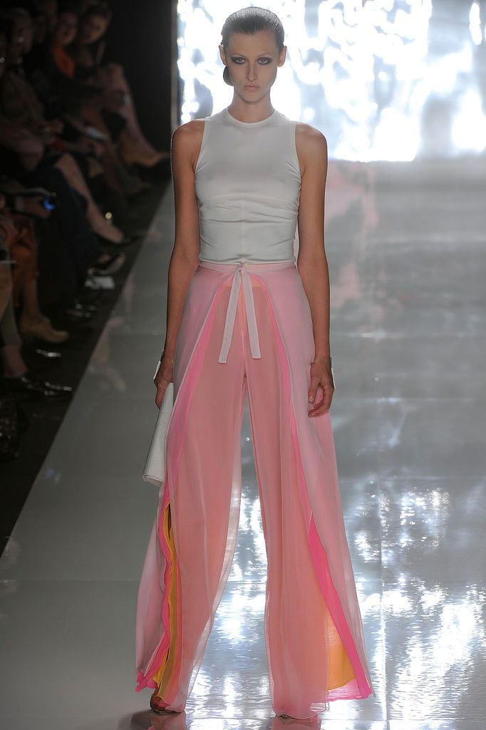 a model walks the runway wearing a white sleeveless top and highwaisted, widelegged pink pants with a tie waist and slits that reveal yellow and orange fabric underneath.