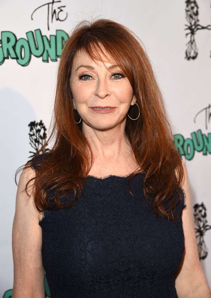 a headshot of a woman with red hair, wearing a black lace dress and smiling. she is at an event and standing in front of a wall with the words "the groundlings" on it.