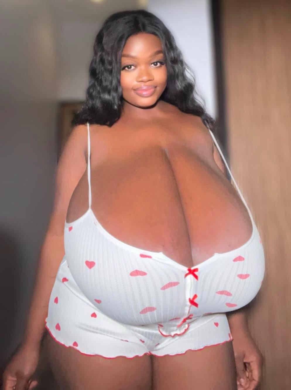 a closeup portrait of a smiling black woman with very large breasts. she is wearing a white pajama set with small red hearts and lips printed on it. the pajama set is a top and shorts. the woman's hair is long and black, styled in loose waves. she is looking directly at the camera. the background is blurry and out of focus.