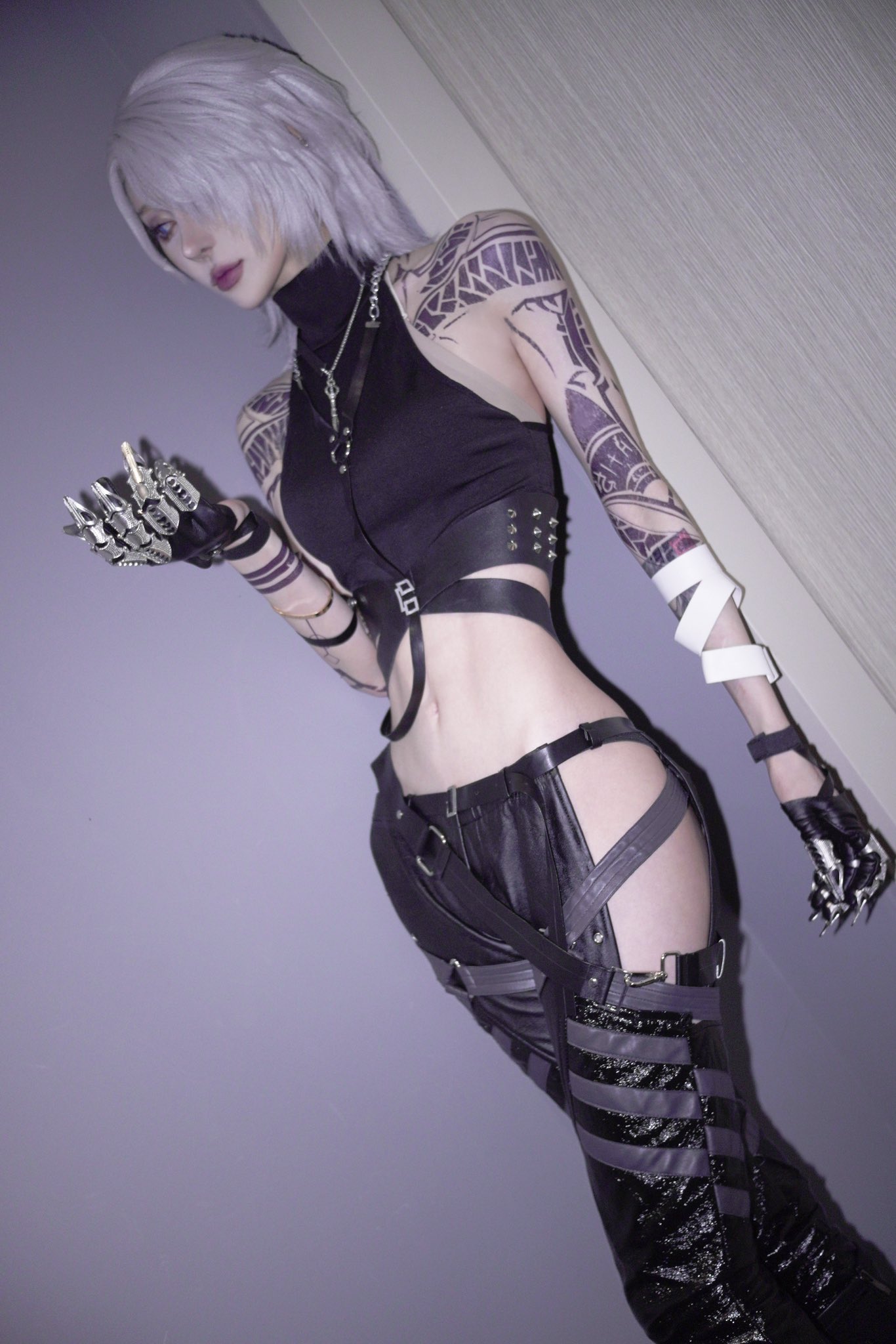 a striking image of a young woman cosplaying a cyberpunkinspired character. she's dressed in a black leather outfit featuring a cropped top, highwaisted pants with straps and buckles, and intricately designed gloves with clawlike accents. her platinum blonde hair is styled in a short, layered cut.  she has detailed tattoos on her arms and a generally lean physique. the background is a muted purplegray wall. the overall mood is dark, edgy, and futuristic.  the image is professionally shot, with good lighting and composition.
