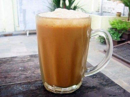 a closeup shot of a glass mug filled with teh tarik, a popular malaysian beverage.  the tea is a rich brown color with a thick, creamy frothy layer on top.  the mug has a clear glass handle and sits on a dark wooden table outdoors. the background is blurred but shows some greenery and a hint of a cafe or restaurant setting. the overall impression is one of a refreshing, flavorful drink enjoyed in a relaxed atmosphere.