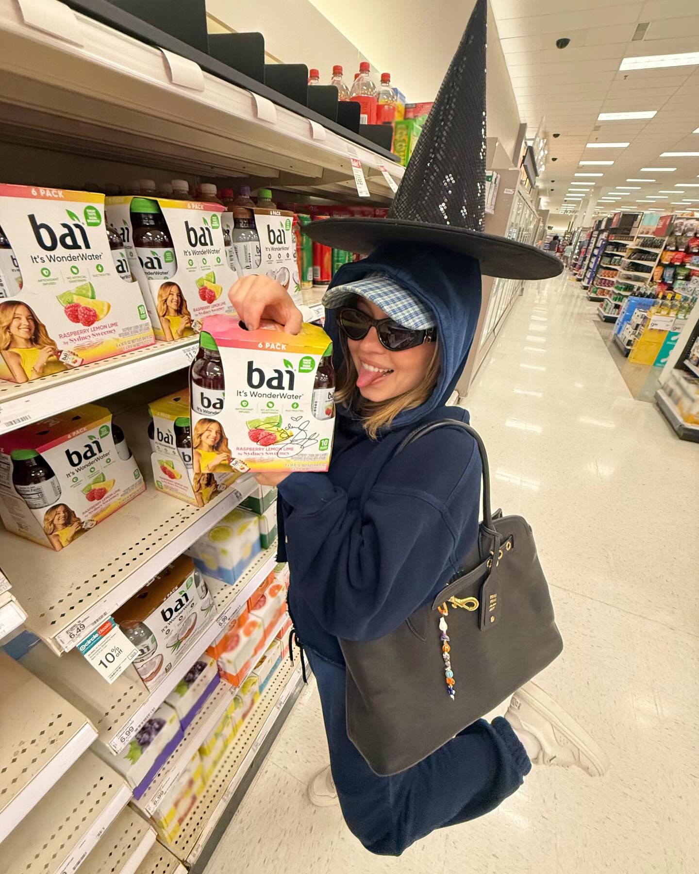 a young woman wearing a witch's hat and sunglasses is kneeling in a target grocery store aisle. she's holding a sixpack of bai raspberry lemonade "wonderwater".  she's wearing a dark blue hoodie and has a darkcolored handbag. several other bai drink boxes are visible on the shelves behind her. the overall mood is casual and fun.