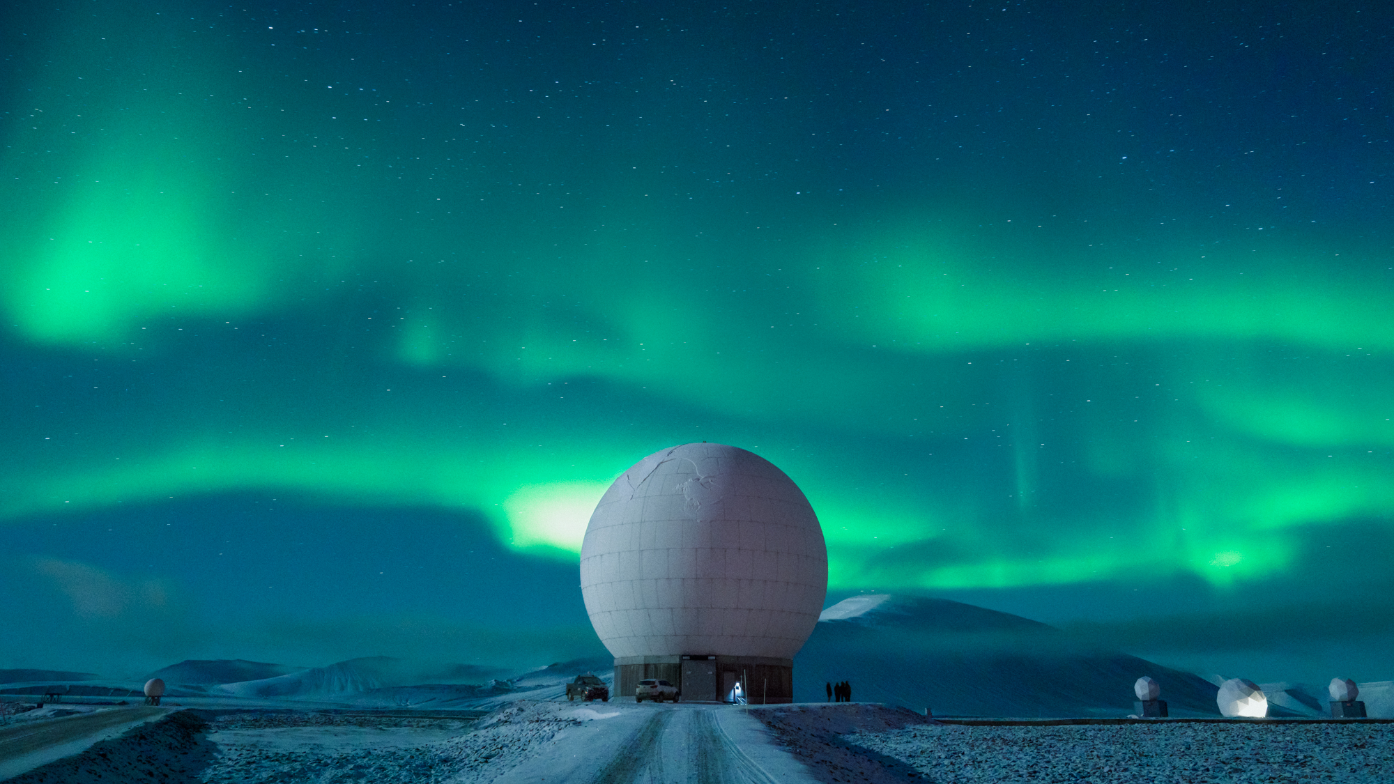 a large white radar dish sits under a vibrant green aurora borealis in a snowy landscape. the aurora fills the night sky, casting an ethereal glow over the scene. in the background, other smaller radar dishes can be seen. the cold arctic climate and the presence of the research facility suggest this is a scientific observatory.